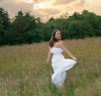 High school senior girl twirling in a white dress in a field at sunset in Newton, Massachusetts – senior portrait by Adriana Kopinja Photography.